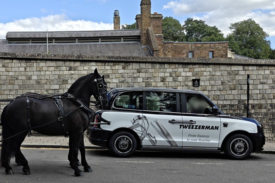 A London black cab with a white Tweezerman superside, parked beside a black carriage horse against a stone wall in Windsor